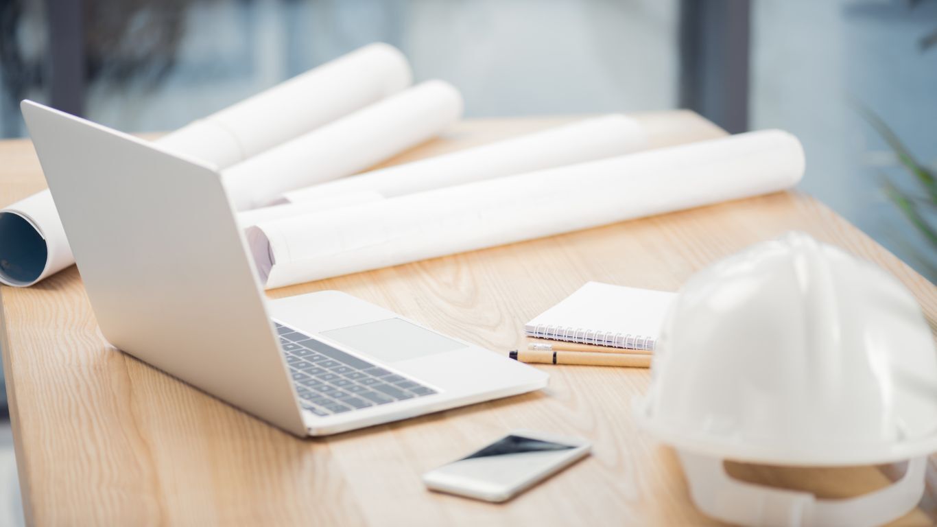 Blueprints, laptop, and hard hat on a desk for balancing field work with contractor planning.