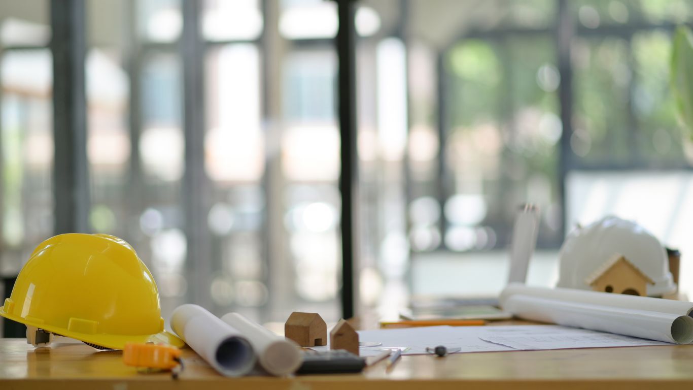 Construction project scheduling tools, blueprints, and a yellow hard hat arranged on an office desk.