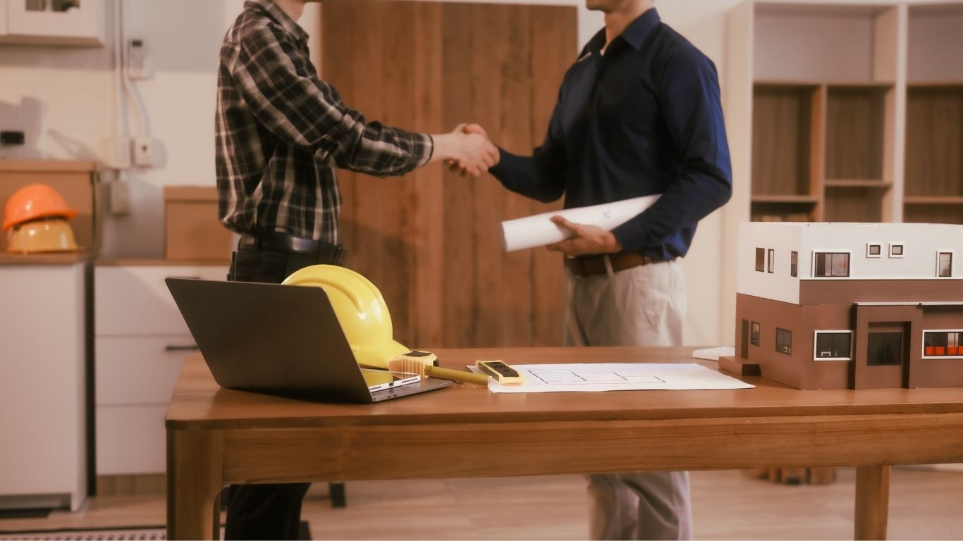 Contractor and homeowner shaking hands beside plans, a laptop, hard hat, and home model after a consultation.